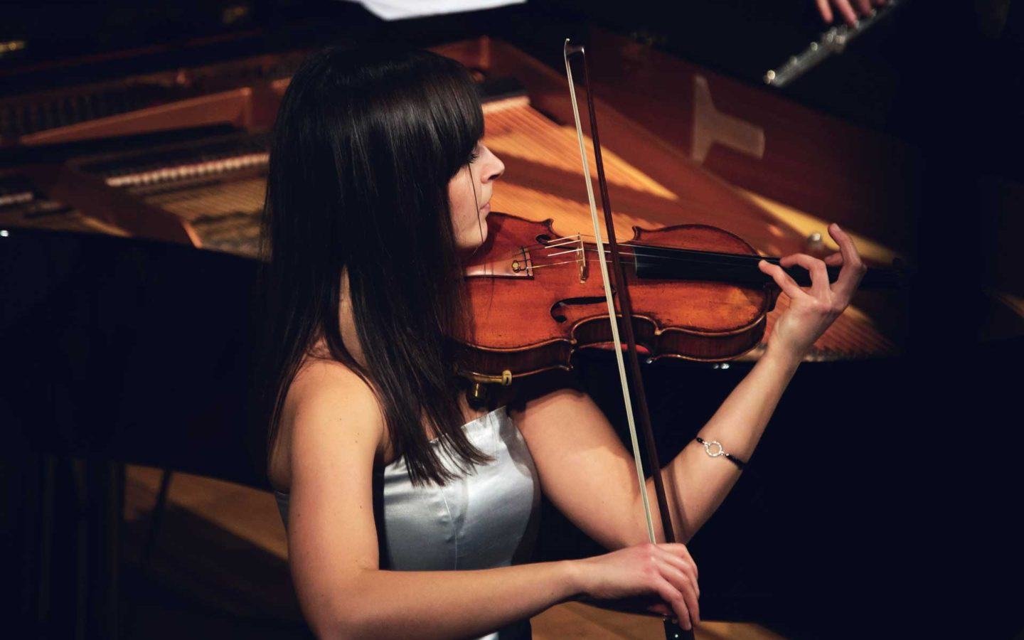 young woman in grey dress playing the violin in front of piano