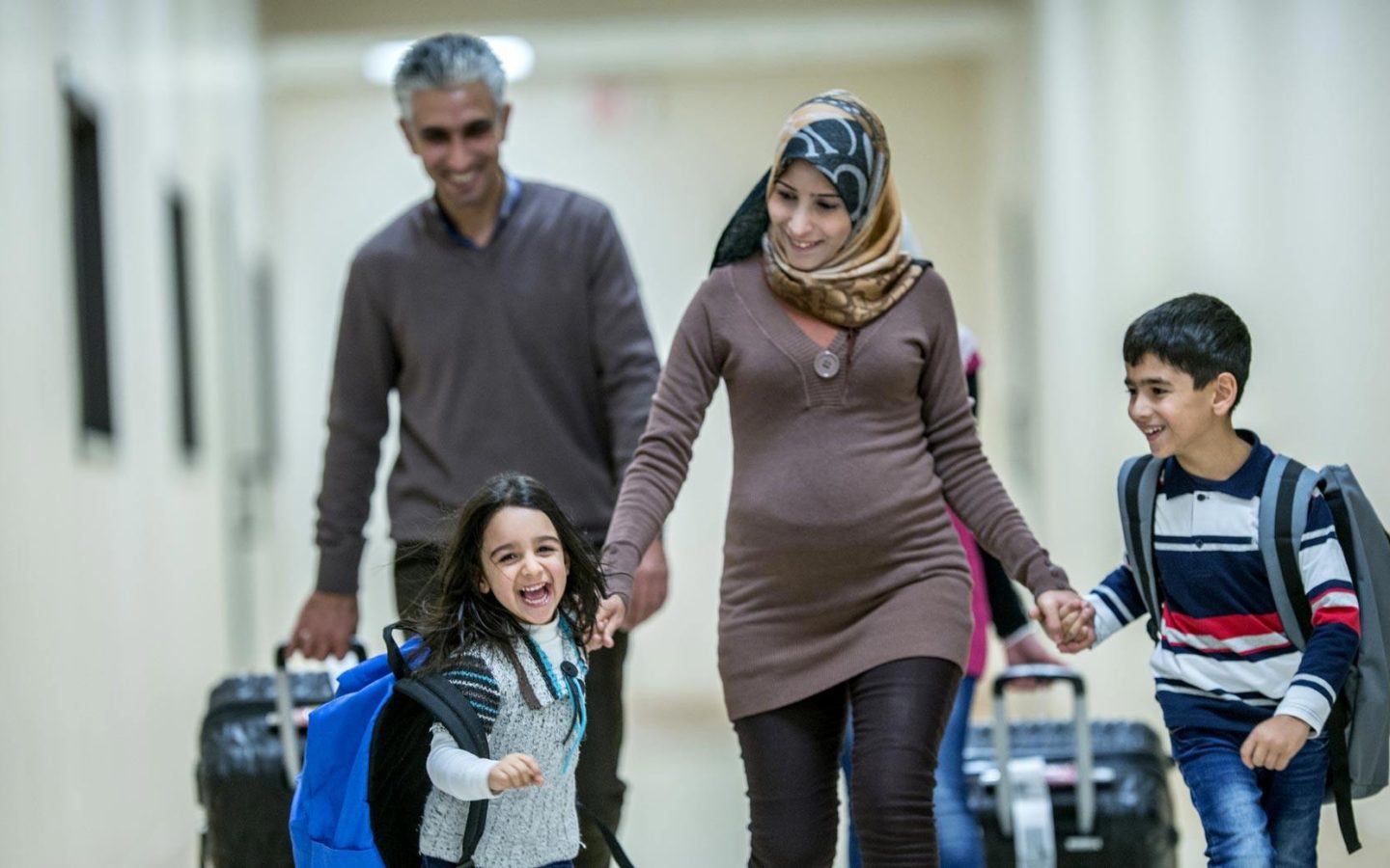 smiling family of four walking down hallway with suitcases and backpacks