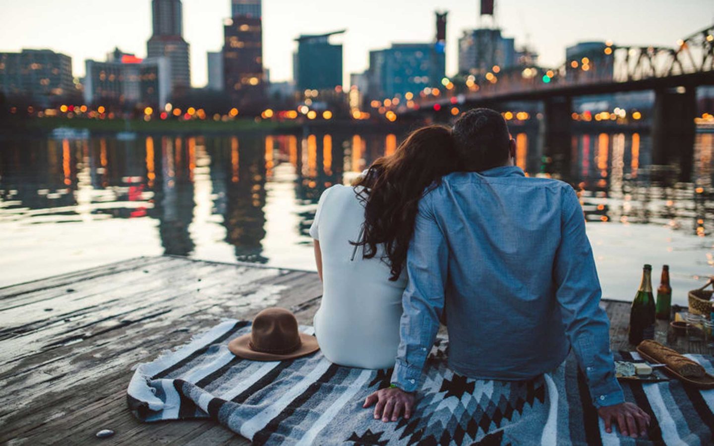 couple sitting on blanket in front of lake with city skyline and bridge in the background